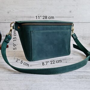 Puede incluir: Un bolso bandolera de cuero verde oscuro con cremallera superior, bolsillo exterior y correa ajustable. Las dimensiones son 28 cm de ancho, 15 cm de alto y 5 cm de profundidad, con una base de 22 cm. Los herrajes son de lat&oacute;n envejecido.