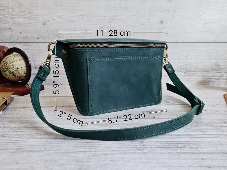 Puede incluir: Un bolso bandolera de cuero verde oscuro con cremallera superior, bolsillo exterior y correa ajustable. Las dimensiones son 28 cm de ancho, 15 cm de alto y 5 cm de profundidad, con una base de 22 cm. Los herrajes son de lat&oacute;n envejecido.