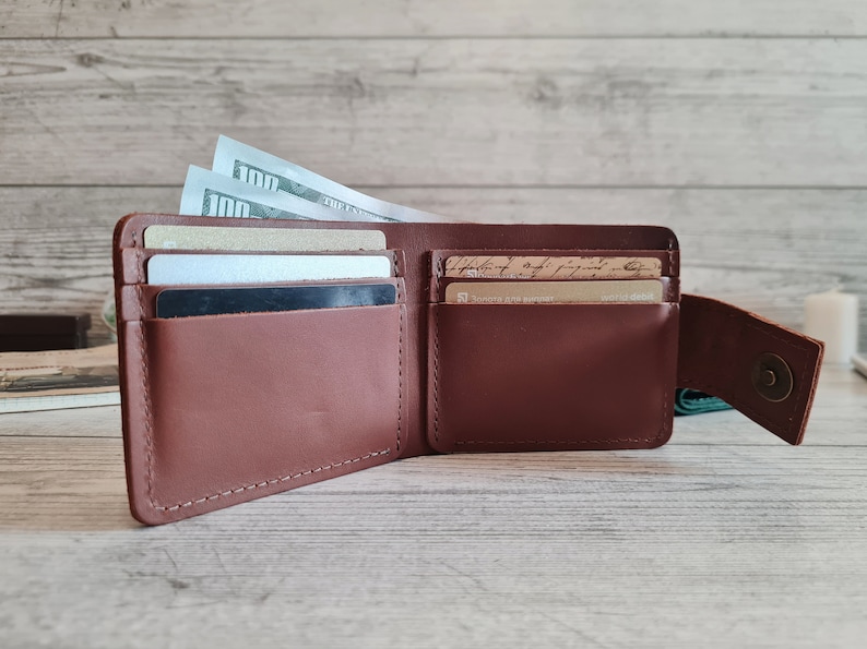 Pu&ograve; includere: Portafoglio in pelle marrone aperto con pi&ugrave; fessure per carte e uno scomparto per contanti. Il portafoglio &egrave; pieno di carte e diverse banconote da cento dollari. Il portafoglio &egrave; aperto, mostrando il suo contenuto.
