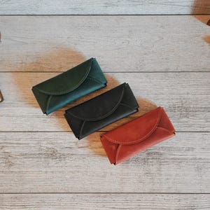 May include: Three leather cases in green, black, and red, arranged on a light wood surface. The cases have a curved flap closure. Other items include a globe, a journal, and a framed picture.