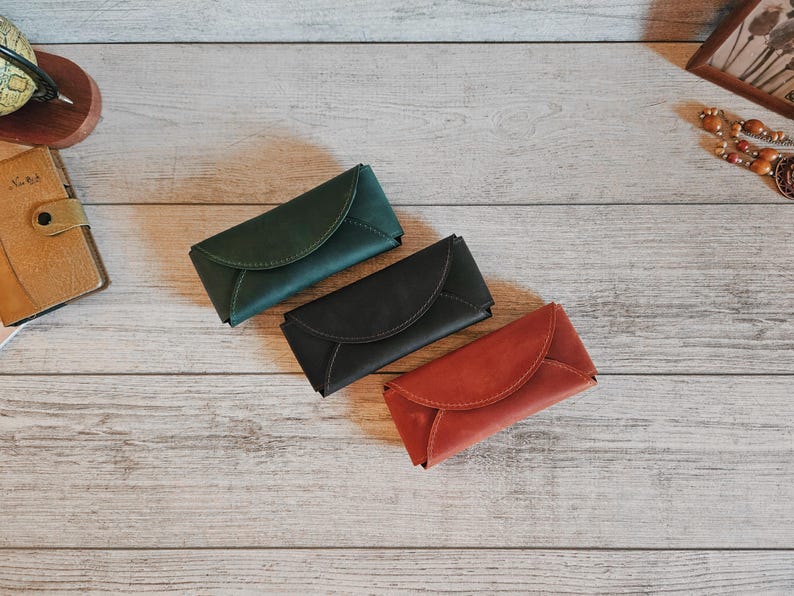 May include: Three leather cases in green, black, and red, arranged on a light wood surface. The cases have a curved flap closure. Other items include a globe, a journal, and a framed picture.