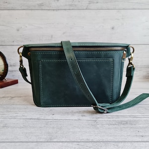 Puede incluir: Un bolso bandolera de cuero verde oscuro con un bolsillo frontal rectangular, costuras visibles, cierre de cremallera superior con herrajes en tono lat&oacute;n y una correa ajustable para el hombro sujeta con clips giratorios de metal.