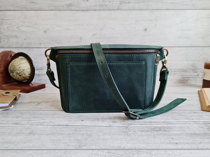 Puede incluir: Un bolso bandolera de cuero verde oscuro con un bolsillo frontal rectangular, costuras visibles, cierre de cremallera superior con herrajes en tono lat&oacute;n y una correa ajustable para el hombro sujeta con clips giratorios de metal.