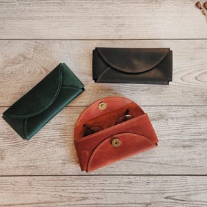 May include: Three leather eyeglass cases in different colors: dark green, dark brown, and red-orange. The red-orange case is open, revealing a pair of glasses. The cases are on a light wood surface.