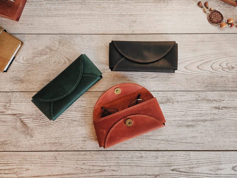 May include: Three leather eyeglass cases in different colors: dark green, dark brown, and red-orange. The red-orange case is open, revealing a pair of glasses. The cases are on a light wood surface.