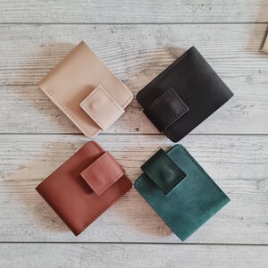 Pu&ograve; includere: Quattro portafogli in pelle beige, nero, marrone e verde acqua sono disposti su una superficie di legno chiaro. Ogni portafoglio ha una chiusura a patta quadrata. I portafogli sono di design minimalista, per carte e contanti.