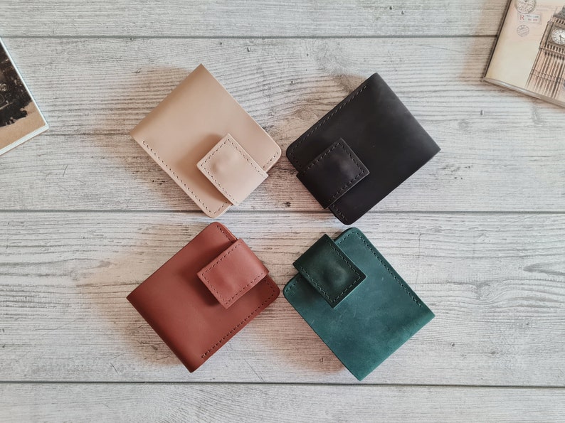 Pu&ograve; includere: Quattro portafogli in pelle beige, nero, marrone e verde acqua sono disposti su una superficie di legno chiaro. Ogni portafoglio ha una chiusura a patta quadrata. I portafogli sono di design minimalista, per carte e contanti.