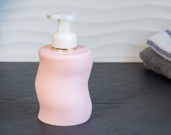 Könnte beinhalten: Ein rosa Seifenspender mit einer weißen Pumpe und einem goldenen Akzent am oberen Rand. Der Spender steht auf einer dunkelgrauen Oberfläche.