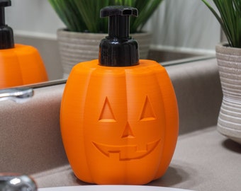 Peut inclure: Distributeur de savon en forme de citrouille orange avec une pompe noire. Le distributeur est posé sur un comptoir de salle de bain blanc.