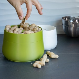 May include: A green and white ceramic peanut dispenser with a hand reaching in to grab a peanut. The dispenser is filled with peanuts and is sitting on a black countertop.