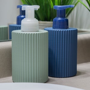 May include: Two decorative soap dispensers with ribbed green and blue bottles and white pump tops. The dispensers are on a light brown countertop.