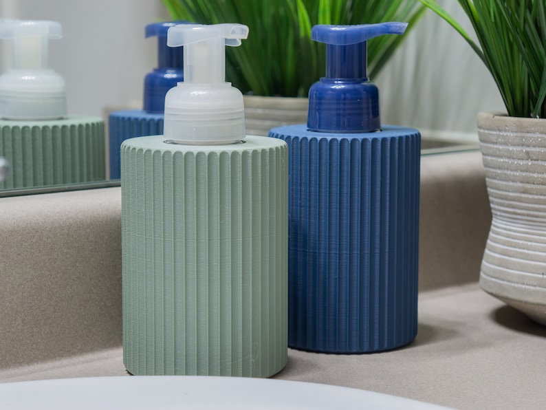 May include: Two decorative soap dispensers with ribbed green and blue bottles and white pump tops. The dispensers are on a light brown countertop.