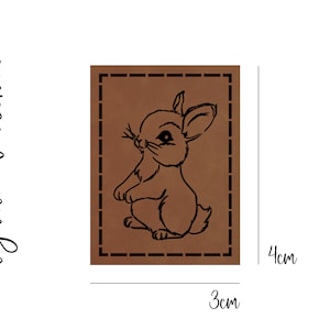 Könnte beinhalten: Ein braunes Lederpatch mit schwarzem Umriss und einem gestickten Rand. Das Patch zeigt eine schwarze Linienzeichnung eines Kaninchens mit großen Ohren und einem flauschigen Schwanz. Das Patch misst 3 cm x 4 cm.