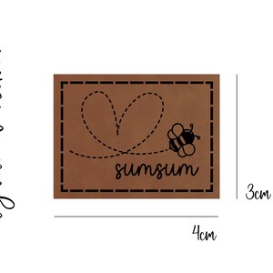 Könnte beinhalten: Braunes Leder-Patch mit einem gestickten Rand. Das Patch zeigt eine schwarze Biene mit einer herzförmigen Linie dahinter. Der Text "sumsum" steht unter der Biene.