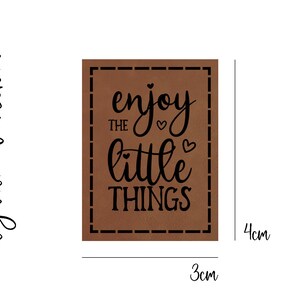 Könnte beinhalten: Braunes Leder-Patch mit einer genähten Umrandung. Der Text "enjoy the little things" ist in schwarzer Kursivschrift mit zwei kleinen Herzen gedruckt.
