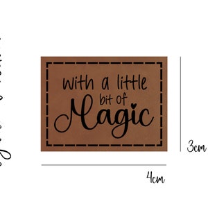 Könnte beinhalten: Ein braunes Leder-Patch mit einem genähten Rand. Das Patch hat den Text "With a little bit of Magic" in schwarzer Kursivschrift.