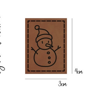 Könnte beinhalten: Ein braunes Leder-Patch mit einer schwarzen Umrandung eines Schneemanns mit Hut und drei Knöpfen. Das Patch misst 3 cm x 4 cm.