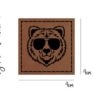 Könnte beinhalten: Ein braunes Lederpatch mit einer schwarzen Silhouette eines Bären mit Sonnenbrille. Das Patch misst 4 cm x 4 cm.