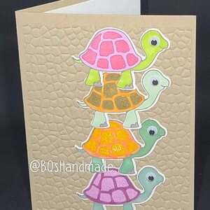 May include: A handmade card with a stack of four colorful turtles on a brown textured background. The card has the text "@BQSHandmade" printed on the bottom left corner.