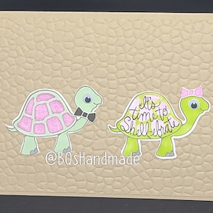 May include: A card with two green turtles on a brown textured background. The female turtle has a pink bow and the male turtle has a black bow tie. The text on the card reads "It's time to Shell-ebrate".