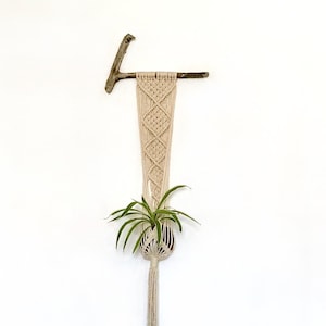 Peut inclure: Un porte-plante en macramé blanc avec une baguette en bois et une plante verte suspendue. Le porte-plante est monté sur un mur blanc à côté d'une porte blanche avec une poignée de porte argentée.
