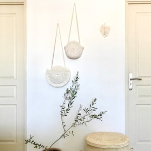 May include: Two white macrame wall hangings with fringe, one larger than the other, are hanging on a white wall. A small white macrame leaf is hanging on the wall to the right of the larger wall hanging. A small potted olive tree is in the lower left corner of the image. A white wicker basket with a lid is in the lower right corner of the image.