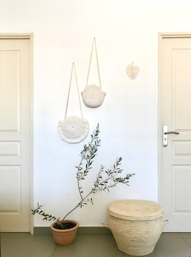 May include: Two white macrame wall hangings with fringe, one larger than the other, are hanging on a white wall. A small white macrame leaf is hanging on the wall to the right of the larger wall hanging. A small potted olive tree is in the lower left corner of the image. A white wicker basket with a lid is in the lower right corner of the image.