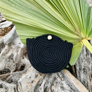 May include: Black round woven bag with a wooden button closure. The bag is sitting on a piece of driftwood with a palm leaf in the background.
