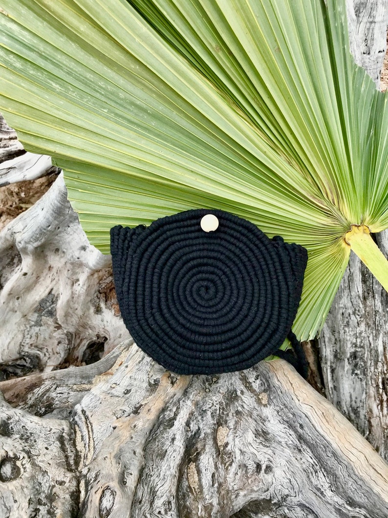 May include: Black round woven bag with a wooden button closure. The bag is sitting on a piece of driftwood with a palm leaf in the background.