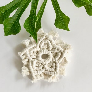 Peut inclure: Une étoile en macramé blanche avec un centre circulaire et des bords frangés. L'étoile est faite de cordon de coton et a un aspect délicat et fait main.