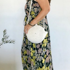 May include: A white round crocheted bag with a long strap worn over a floral maxi dress with a black background and yellow, pink, and white flowers.