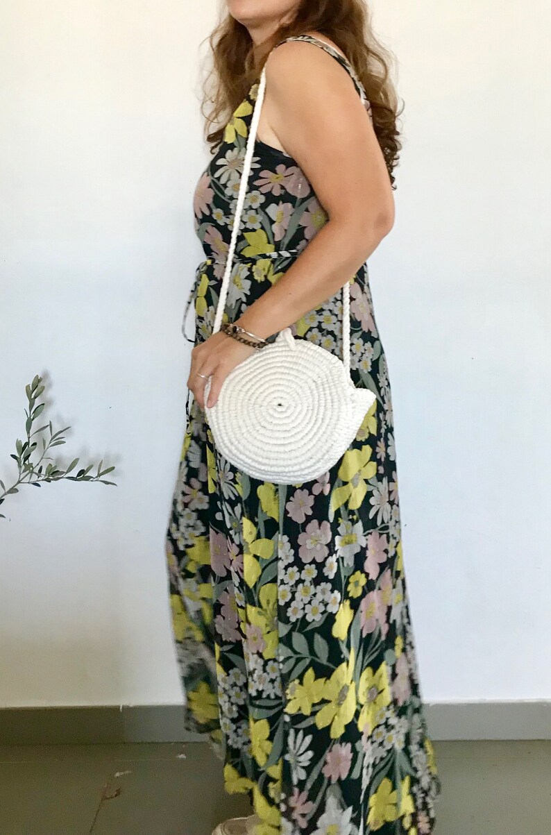 May include: A white round crocheted bag with a long strap worn over a floral maxi dress with a black background and yellow, pink, and white flowers.
