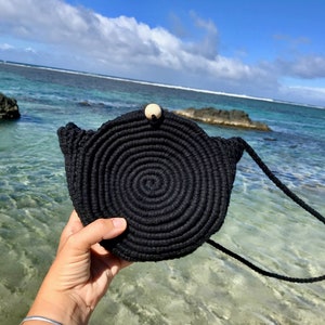 May include: A black round crocheted bag with a wooden button closure and a long black strap. The bag is held in a hand against a background of blue water and a cloudy sky.
