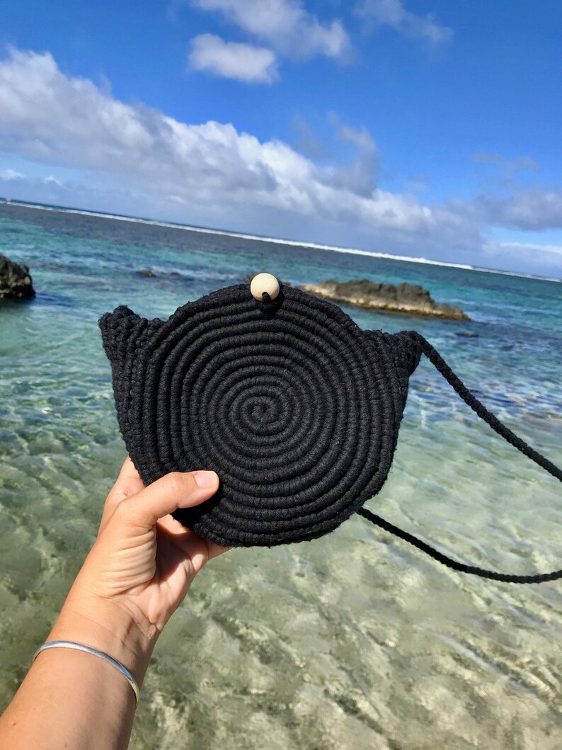 May include: A black round crocheted bag with a wooden button closure and a long black strap. The bag is held in a hand against a background of blue water and a cloudy sky.