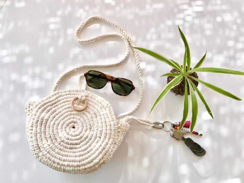 May include: A white crocheted round bag with a wooden ring handle and a long white rope strap. The bag is lying on a white surface with a pair of brown sunglasses and a potted plant.