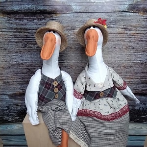 May include: Two plush goose figurines wearing straw hats and clothing. The goose on the left is wearing a plaid vest and a plaid skirt. The goose on the right is wearing a floral dress with a plaid skirt and a red bow.