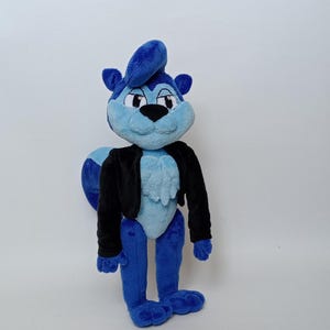 Pode incluir: Um peluche de esquilo com um corpo azul vibrante, rosto e peito num azul mais claro. A personagem tem uma madeixa de cabelo azul na cabeça, uma cauda grande e felpuda, e veste um casaco preto de manga comprida sobre o pelo azul macio.