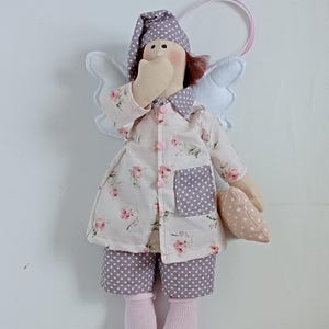 May include: A handmade angel doll wearing a floral print nightgown, polka dot shorts, and a matching nightcap. The doll has white wings, pink socks, and is holding a small pillow. A pink ribbon is attached for hanging.