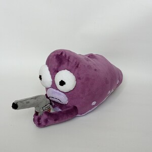 Puede incluir: Peluche morado que representa una criatura alien&iacute;gena con grandes ojos blancos y una pistola de juguete gris. El peluche tiene una textura suave y difusa y est&aacute; sobre un fondo blanco liso. La boca de la criatura es de color morado claro.