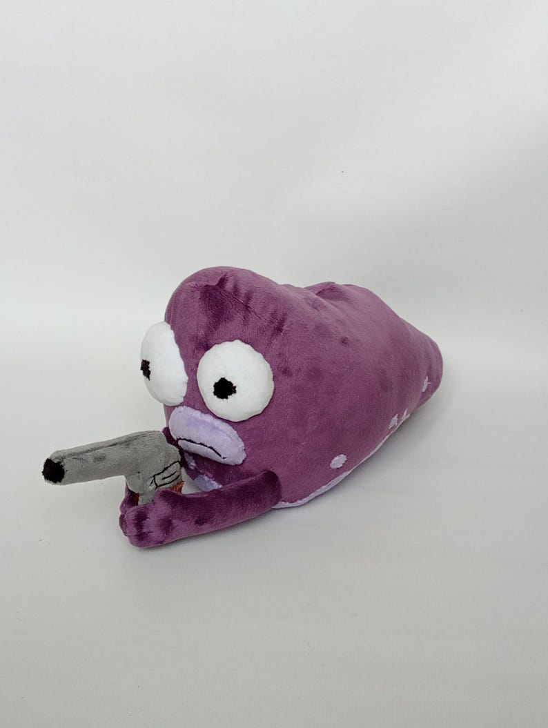 Puede incluir: Peluche morado que representa una criatura alien&iacute;gena con grandes ojos blancos y una pistola de juguete gris. El peluche tiene una textura suave y difusa y est&aacute; sobre un fondo blanco liso. La boca de la criatura es de color morado claro.