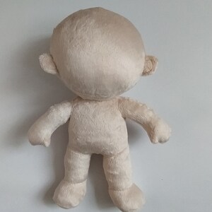 Op de afbeelding: Een beige pluche pop met een rond hoofd, kleine oren en zonder gezichtskenmerken. De pop heeft armen en benen en staat op een wit oppervlak.