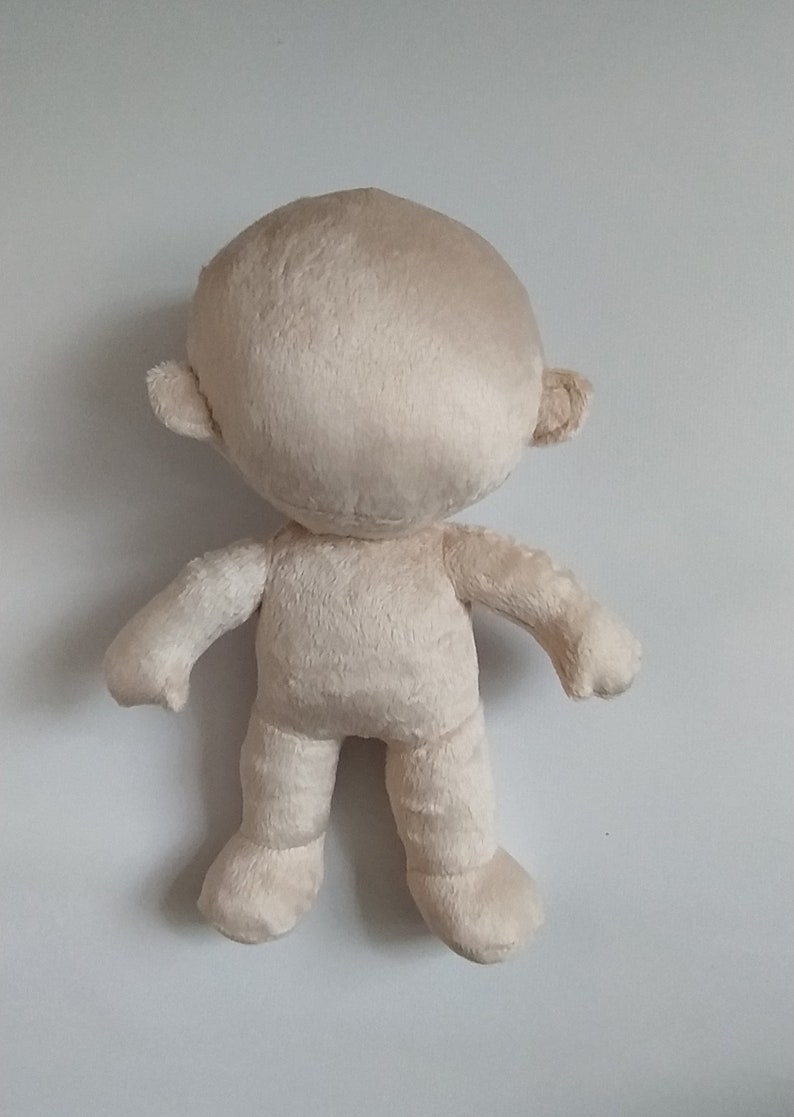 Op de afbeelding: Een beige pluche pop met een rond hoofd, kleine oren en zonder gezichtskenmerken. De pop heeft armen en benen en staat op een wit oppervlak.