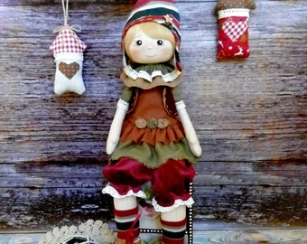 Pode incluir: Uma boneca feita à mão com um chapéu listrado vermelho e verde, um vestido castanho com saia verde e meias listradas vermelho e branco. A boneca está sentada numa caixa de madeira com uma renda branca e uma decoração em forma de estrela branca à sua frente.