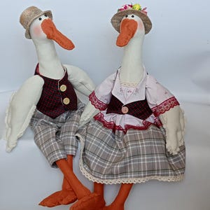 May include: Two plush goose figurines, one wearing a plaid skirt and a pink blouse with lace trim, the other wearing a plaid vest and pants. Both have orange beaks and feet and are wearing hats.