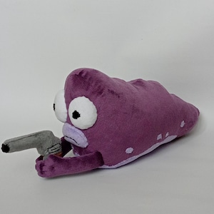 Puede incluir: Peluche con forma de alien&iacute;gena morado con grandes ojos blancos y una pistola de juguete gris. El alien&iacute;gena tiene una boca de color p&uacute;rpura claro y peque&ntilde;os puntos blancos en su cuerpo. El juguete est&aacute; hecho de material suave.
