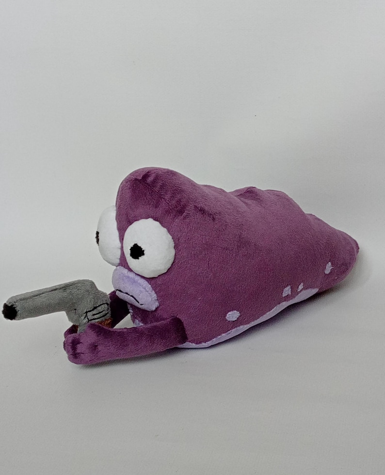 Puede incluir: Peluche con forma de alien&iacute;gena morado con grandes ojos blancos y una pistola de juguete gris. El alien&iacute;gena tiene una boca de color p&uacute;rpura claro y peque&ntilde;os puntos blancos en su cuerpo. El juguete est&aacute; hecho de material suave.