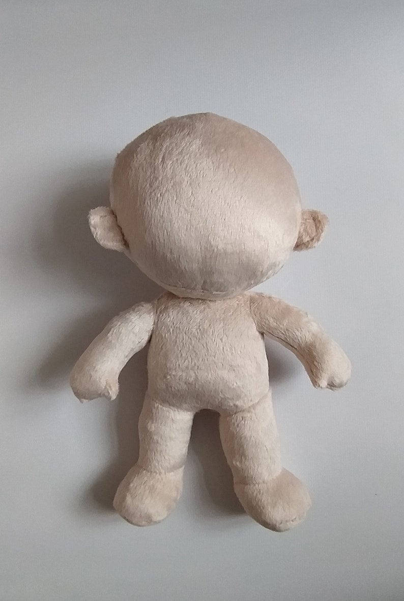 Op de afbeelding: Een beige pluche pop met een rond hoofd, kleine oren en een eenvoudig lichaam. De pop staat op een wit oppervlak.