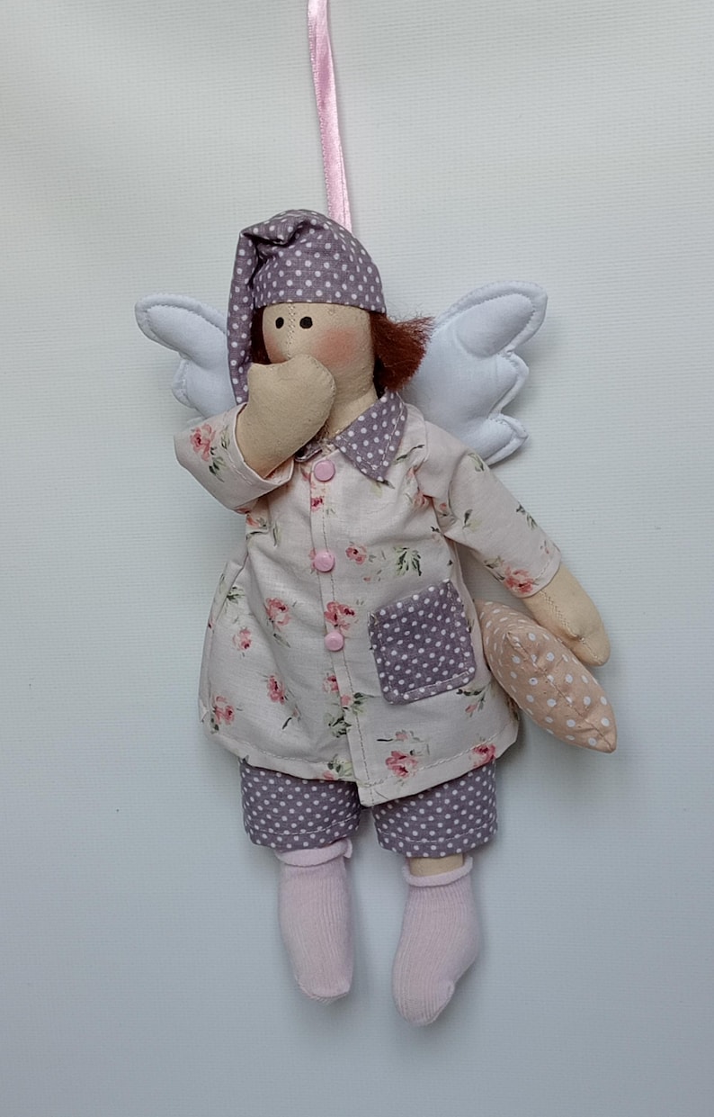 May include: A handmade doll dressed in pajamas with a floral print and polka dot accents. The doll has white wings, a nightcap, and is holding a small pillow. The doll is wearing pink socks and is suspended by a pink ribbon.