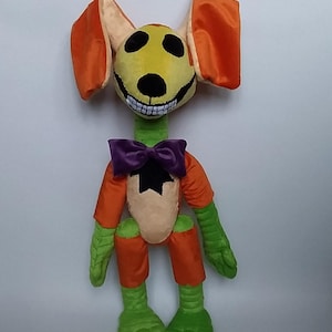 May include: A plush toy creature with a yellow face, orange ears, and a wide, toothy grin. It has a green neck, arms, and legs, and wears orange shorts and sleeves. A purple bow tie and a black star adorn its chest. The toy is designed to be a whimsical character.