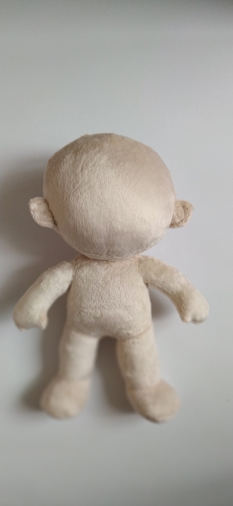 Op de afbeelding: Een beige pluche pop met een rond hoofd en zonder gezichtskenmerken. De pop heeft kleine oren en staat op een wit oppervlak.